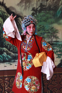 Chinese Peking Opera performer poses in a bright red outfit, carrying a box wrapped in a yellow cloth.