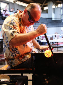 Glass artist David Graeber in a hawaiin shirt sitting at a glass studio bench turning a glowing hot paperweight at the end of a steel rod.