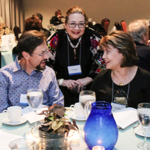 Two people look at each other smiling at a set table. Behind them a woman smiles, looking at the camera.