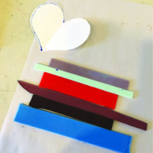 Strips of glass in multiple colors sit next to a cut white paper heart.