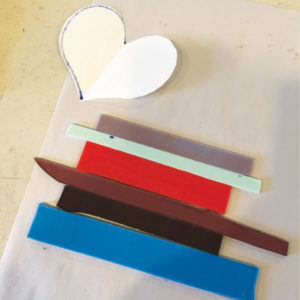 Strips of glass in multiple colors sit next to a cut white paper heart.