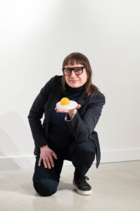 Glass Artist Deb Czeresko smiles and kneels, holding a blown glass egg. Photo credit Paula Bernadette Robinson.