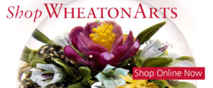 Shop WheatonArts banner with red text. The background image is a closeup of a paperweight by Paul Stankard with a rich magenta flower with a yellow center and green leaves.