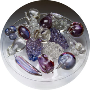 A circle cutout of Dish in Tuning Color by Alessandro Moretti. It is a large clear glass plate with purple and clear fruit on it, like apples, grapes, and cherries.