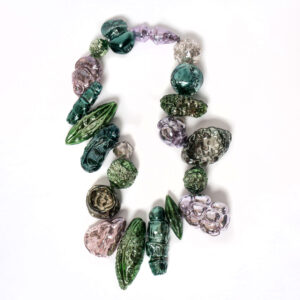 Image of a glass piece on a white background. The piece is shaped like an oval that bends in at the sides. It is comprised off alternating green, dark blue green, light pink, and light purple glass pieces of various shapes and sizes. The piece resembles a beaded glass necklace.