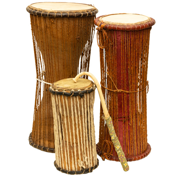 Image of three pieces from the Ceremonies in Circles exhibit on a white background. There are two tall drums that are wide at the ends and thin in the middles. The drum on the left has a light brown base with light brown stings extended vertically from the top of the drum to the bottom. A white strand is tied around the middle. The drum on the right is slightly shorter than the one on the left with a light brown strand tied around the middle and a reddish base color. In front of them is a smaller drum about half the size of the taller ones. This drum has a light beige base color and white vertical strands. Resting on top of the drum is the curved portion of a long, thin light beige piece with green patterned fabric wrapped around the end.