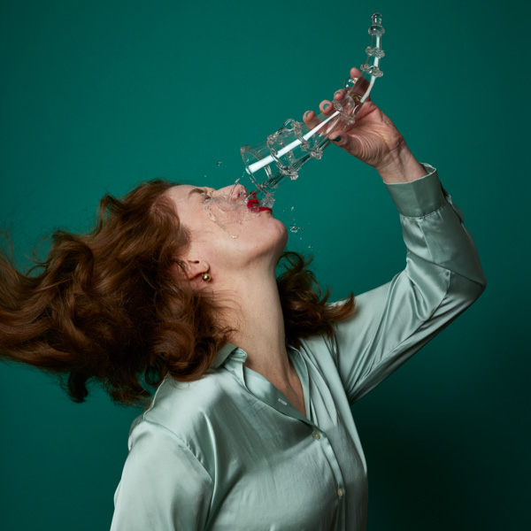 Image of a lady with a clear horn-shaped trick glass in front of a dark teal background. The lady has her head tilted back as she pours a clear liquid out of the trick glass into her mouth. The liquid instead spills all over her face ,mouth, and light teal colored shirt. The lady has shoulder length, curly brown hair and red lipstick.
