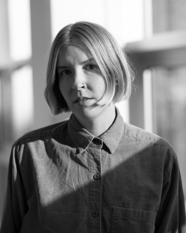 Black and white headshot image of WheatonArts 2024 Creative Glass Fellow: Brianna Gluszak. Brianna has short hair that is cut above the chin and is wearing a long sleeve shirt with buttons. a collar, and a pocket on the right side.
