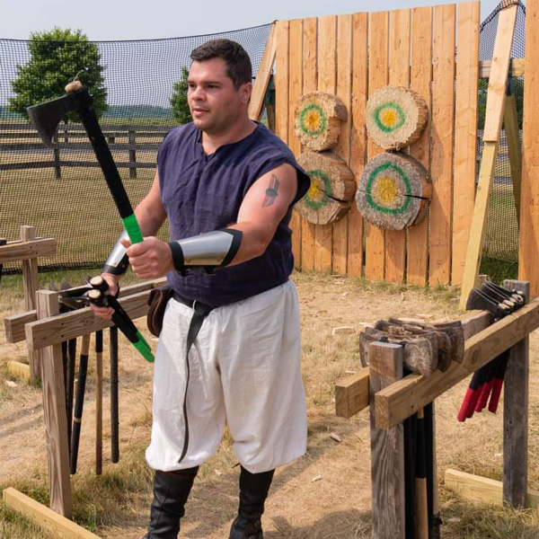 Image of a Fantasy Faire performer outside dressed in a black tank top, white baggy pants, a black belt, black high boots, and silver and black bracers on each wrist. One hand is holding an axe with a green handle in a throwing position and the other hand is holding two more by the waist. More axes rest on wooden stands on both sides of the performer. There are wooden planks standing in the background like a wall with four circular slabs of wood with green and yellow circles painted onto them as targets. The circles are arranged in a square formation in the center of the wall. There is black netting around the wood and the ground is sandy.