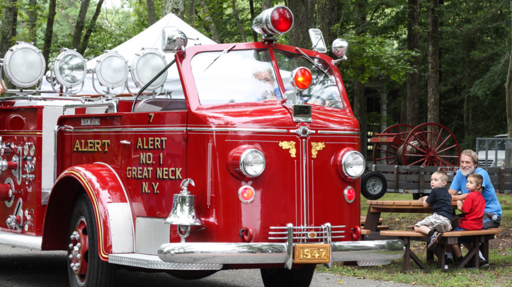 Image of a red antique fire truck outside at WheatonArts during Fire Muster. The fire truck is being driven on the pavement while two children and an adult sit at a brown picnic table to watch the truck drive by.