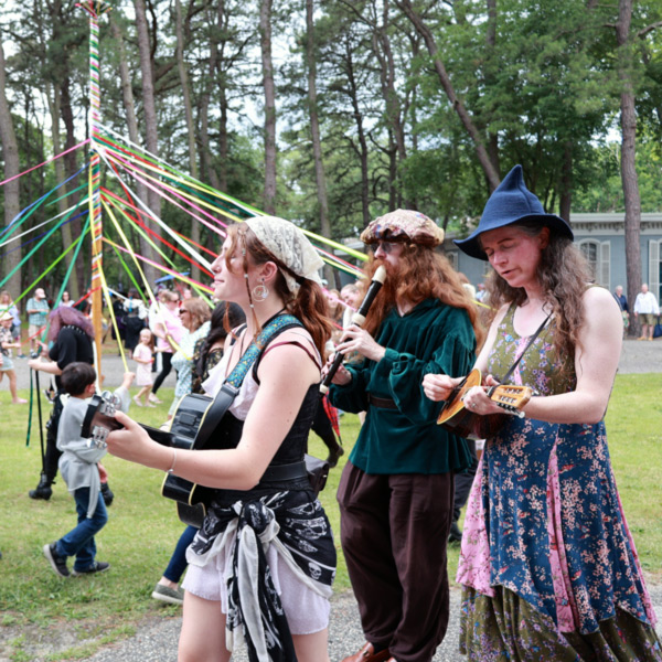 Image of three performers outside at Fantasy Faire at WheatonArts. Each performer is dressed in their own unique fantasy garb and is playing an instrument. Two of the performers have string instruments and one has a flute-like instrument. In the background are Fantasy Faire visitors participating in the rising of the Spring Pole. The pole is tall, thing, and a light brown color. The visitors are wrapping colorful ribbon around it.