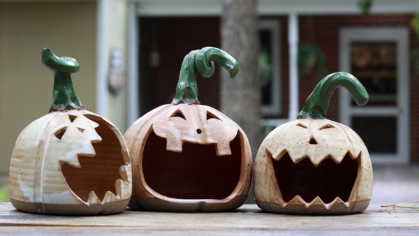 Image of three clay jack-o-lanterns lined up in a row outside in front of the WheatonArts Pottery Studio. The jack-o-lanterns have large open mouths, curly green stems, and a beige pumpkin body.