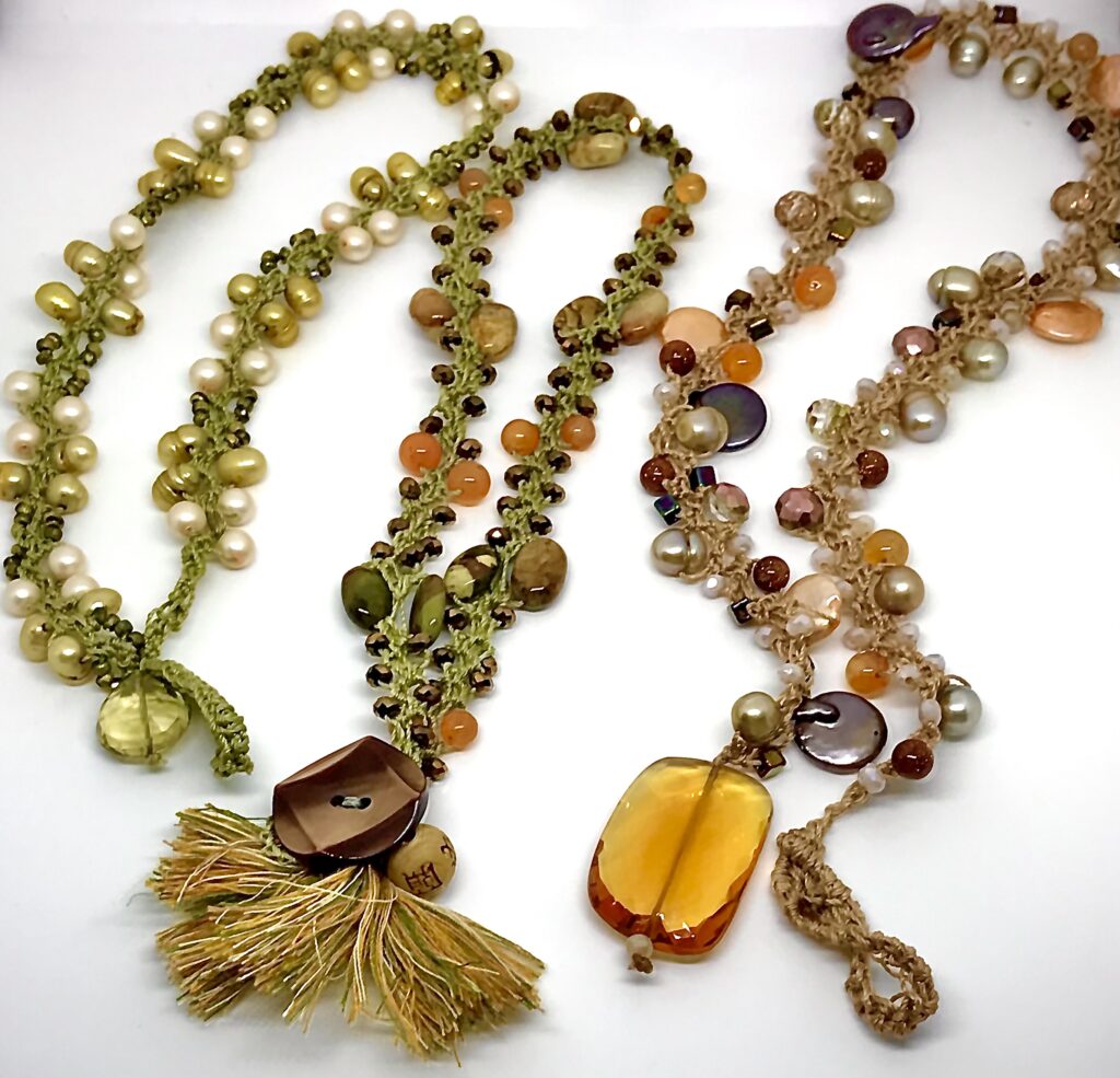 Image of two necklaces. The necklace on the left has a large brown bead and light green and beige fringe. There is a small round beige bead to the left of the brown bead. The chain is green with orange, green, orange, and white beads on each side in sets of two. There are even smaller brown beads in between the sets of two. The necklace on the left has a yellow rectangular bead with red at the bottom. The chain is beige with round silver, gold, brown, orange, beige, and pink beads as well as some flat beads throughout the cord.