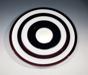 Image of a large glass plate against a white to dark gray gradient background. The plate has alternating black and white circular stripes until it reaches the center. There are two black and two white stripes. The center has a thick black stripe around a white circle.