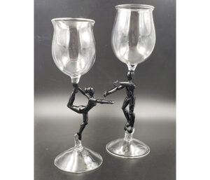 Image of two glass goblets in front of a gray background. The goblets are clear at the top and bottom. The stem of the glass on the right is comprised of a dark gray woman standing on one leg with an arm extended in front of her. Her other arm and leg are holding the top of he glass. The goblet on the right has a stem that is comprised of a dark gray man with both arms extended in front of him while the top of the glass is balanced on his head. His knees are slightly bent.