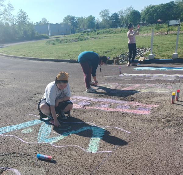 Image of three people outside in the WheatonArts circular parking lot. Two people are bent over to use cans of spray chalk to create a colorful walkway across the pavement to where the center garden section of circle is. The third person towards the back of the image is standing and shaking a can of the spray. Each line of the walkway has a unique design.