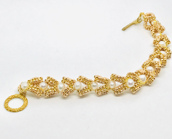 Image of an unhooked bracelet on a white backdrop. The bracelet is comprised of alternating small white spherical beads and "v"-shaped clusters of small gold spherical beads. There is a circular gold attachment on the left side of the bracelet and a small stick-like gold piece on the right.