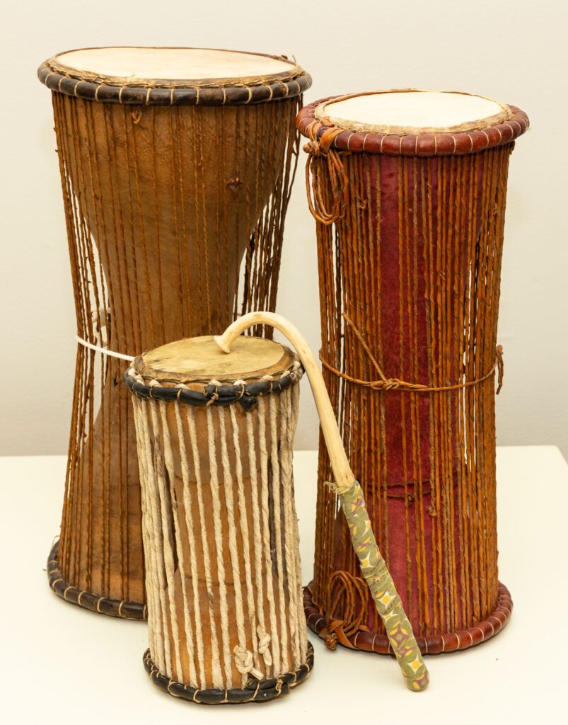 Image of three pieces from the Ceremonies in Circles exhibit on a white background. There are two tall drums that are wide at the ends and thin in the middles. The drum on the left has a light brown base with light brown stings extended vertically from the top of the drum to the bottom. A white strand is tied around the middle. The drum on the right is slightly shorter than the one on the left with a light brown strand tied around the middle and a reddish base color. In front of them is a smaller drum about half the size of the taller ones. This drum has a light beige base color and white vertical strands. Resting on top of the drum is the curved portion of a long, thin light beige piece with green patterned fabric wrapped around the end.