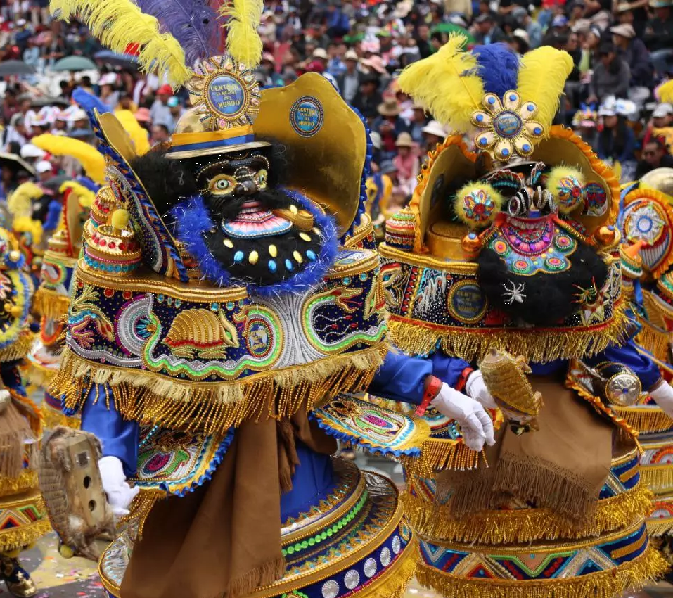 Image of two people dressed in vibrant traditional clothing and masks participating in a Morenada Mask Dance with a large crowd in the background.