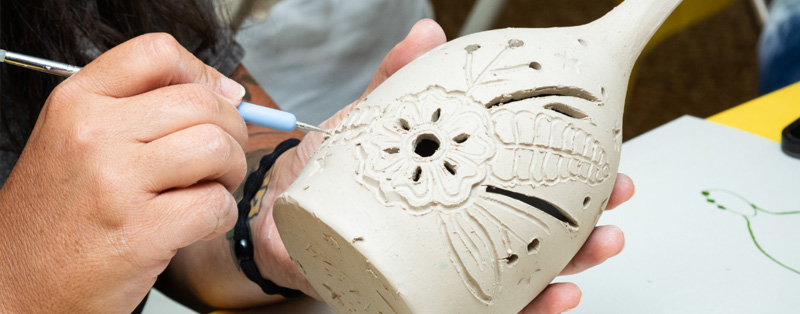 Close up image of a white wet clay luminary being crafted. The luminary is cylindrical with a tall, thin bottle neck-like piece at the top. The luminary is being held with one hand and the other is using a sculpting tool to draw and carve a floral design into the clay.