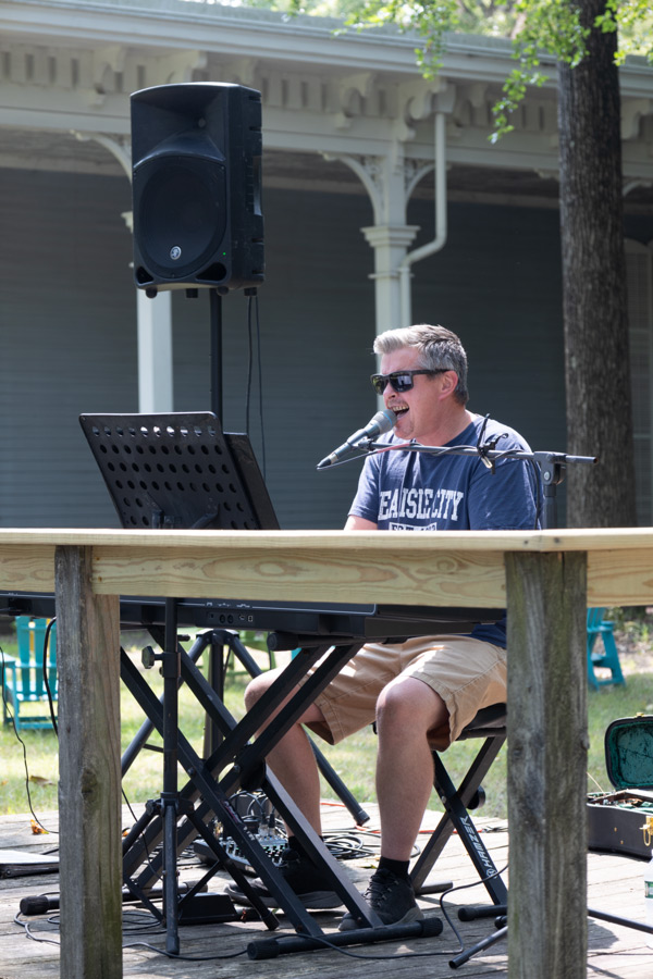 Image of Dale Bierman sitting outside on a wooden stage next to the WheatonArts Museum of American Glass. Dale is singing into a microphone and is using both hands to play the large keyboard that is in front of him. There is a tall speaker in the background on the left side of the image.