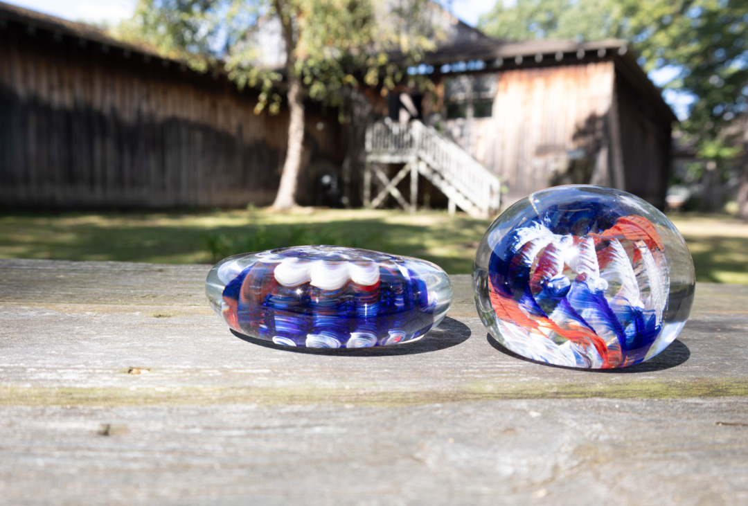 An image of two red, white, and blue swirl glass paperweights resting outside on a wooden picnic table in front of the WheatonArts Glass Studio. The paperweight on the left is disc-shaped, while the paperweight on the right is orb-shaped. They both are on the right side of the image.