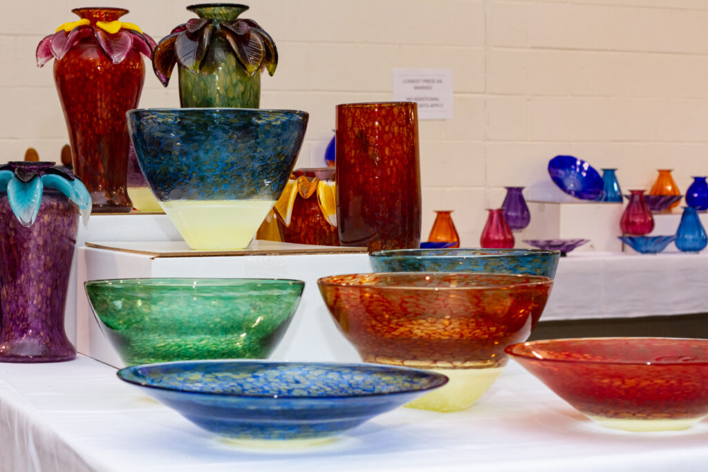 Image of glass bowls and vases from David and Melanie Leppla. There are five bowls displayed on a white tablecloth-covered table. The bowls have beige bottoms with either a red, blue, or green top. Each bowl has a different height and width. There is also a tall cylindrical red vase and three more tall vases with floral glass petals around the neck. One of the vases is red with dark pink petals with smaller yellow petals on top. Another vase is green with dark green and brown petals. The final vase is purple with light blue petals with smaller purple petals on top of them. There are more vases and bowls displayed in the background.