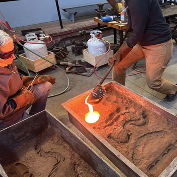 Image of an artist inside of the WheatonArts Glass Studio using a large rod with a scoop on the end to pour hot glass into a large mold with a swirling shape carved into it while another artist is ready to assist