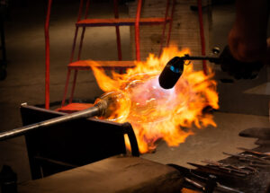 Close up image of a torch being used on a mass of hot glass at the end of a metal rod. The hot glass has been lit up in orange flames that extend all around the glass. The flames are a pinkish red around the area the torch is aiming.