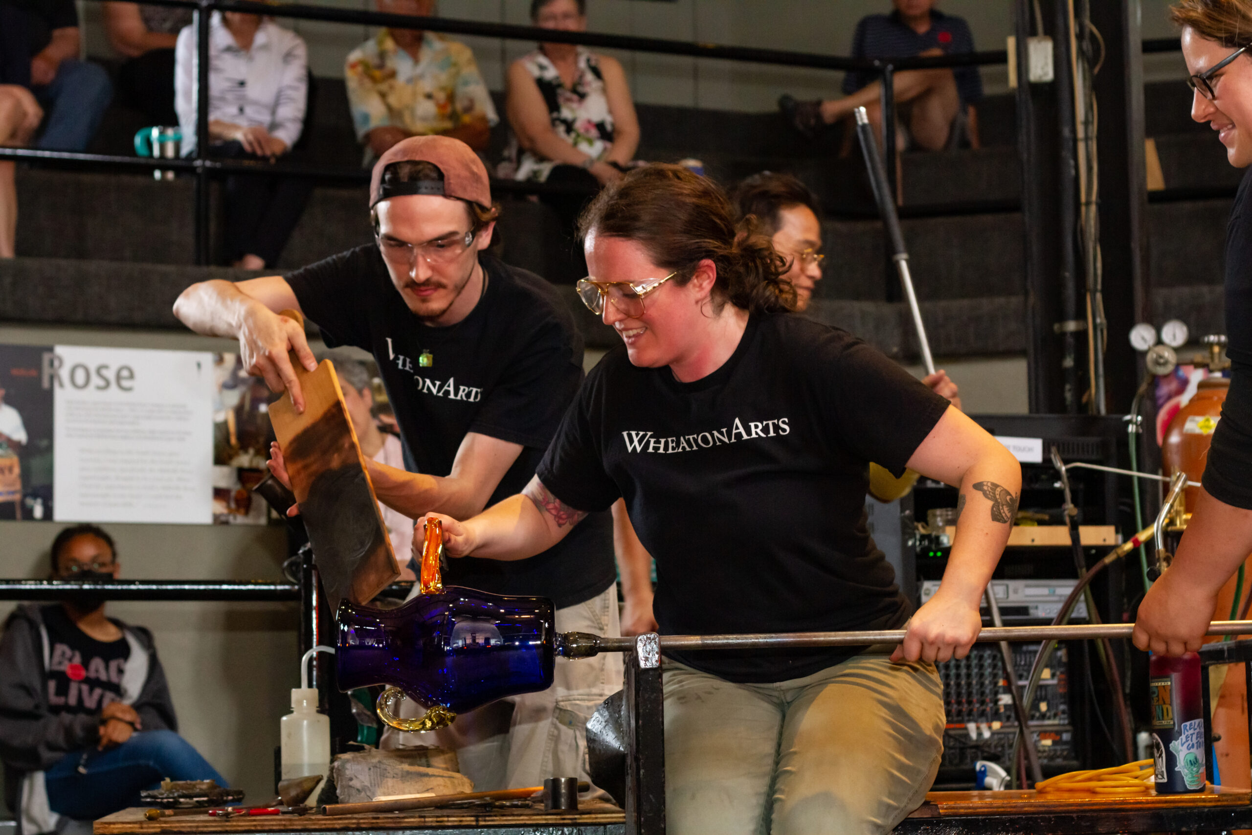 Image of four members of the WheatonArts Glass Studio Team working inside of the WheatonArts Glass Studio in front of a crowd of people. One member is sitting at a glassblowing bench with a long metal rod with a large cobalt blue vase at the end of it. One side of the vase already has a clear handle that is slightly orange from the heat, The team member is working on forming the handle on the other side of the vase. The glass that is being shaped is bright orange from the heat. The same team member also has a hand on the metal rod and is being assistant by the hands of a second team member, slightly off camera on the right side of the image. A third team member is using both hands on a large wooden paddled to help shape the handle. A fourth team member can be seen working in the background. Each team member is wearing clear goggles and a black t-shirt with white text that reads "WheatonArts".