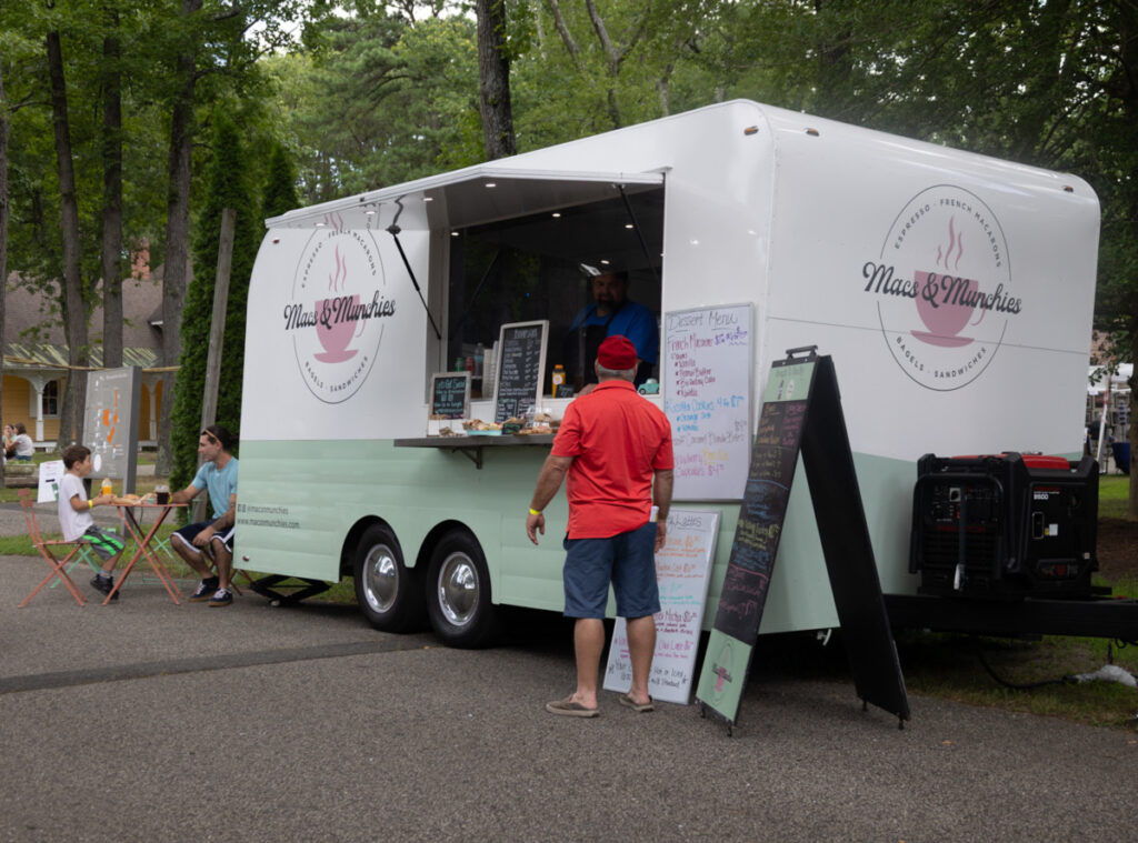 Image of the white and mint colored Mac & Munchies food truck outside at an event at WheatonArts. There are two white boards and three chalkboards featuring their menu. There is a customer standing in front of the window and two more sitting at a table and chair set up on the left side of the truck.