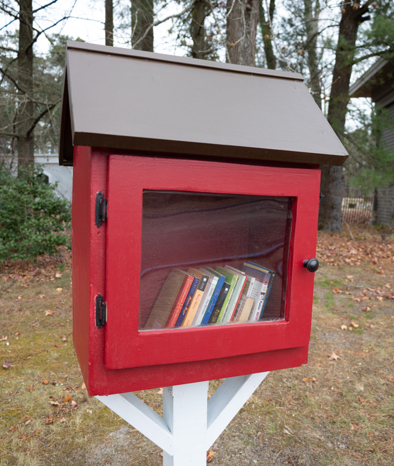 Image of the Little Free Library that is outside on the WheatonArts grounds. The library is square shaped and red with a dark brown triangular roof. The hinges and knob of the red door are black and there is a clear window on the door to see the books inside. The library rests on top of a white stand.