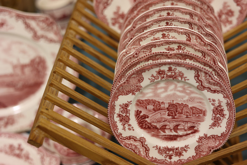 Image of the banner for the 2025 WheatonArts Mid-Winter Antique Show. The background image is blurred, but the right side of the image is in focus and shows a close-up of a brown rack of red and white antique plates with detailed floral designs around a landscape in the center.