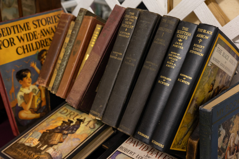 Close-up image of 15 antique books displayed at the WheatonArts Mid-Winter Antique Show. 13 of them are lined up on top of the other two. Visible titles include "Bedtime Stories for Wide-Awake Children", "Drums", "The Children of the New Forest", "The Story of Siegfried", "The Scottish Chiefs", and "The Black Arrow".