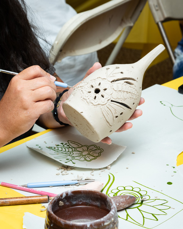 Close-up image of a white clay luminary being worked on over a craft table. One hand is holding the luminary while the other is using a light blue tool to carve a floral design into the soft clay. Floral designs have been drawn onto papers that are scattered onto the craft table beneath them. Also on the table are more tools and a brown clay mug that is filled with water.