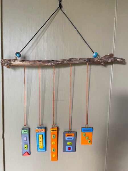 Image of five fused glass rectangles of various lengths and widths are hanging with long orange string from a brown wooden branch. The branch is hanging with long black string on a white wall from a nail. There is a small blue bead on each side of the black string where it the ends meet the branch. The fused glass pieces are gray, blue, and orange at the base with small colorful rectangular and square attachments fused on top of them for decoration.