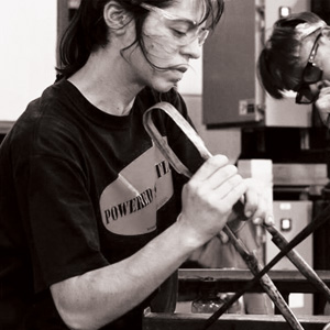 Grayscale side profile image of Anna Antonia Yiaxi Savvidou working at a glassblowing bench. Anna is using both hands to shape hot glass with a large pair of metal tweezers. A small corner of the hot glass is pictured at the bottom right of the image.