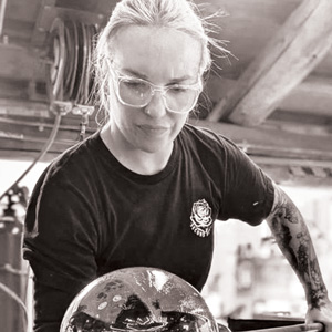 Grayscale image of Danielle Brensinger. The image shows Danielle sitting at a glassblowing bench with a metal rod across the the sides of the bench. The top of a large piece of hot glass is pictured in the image at the end of the metal rod. Danielle has light hair, is wearing glasses, and is looking at the hot glass.