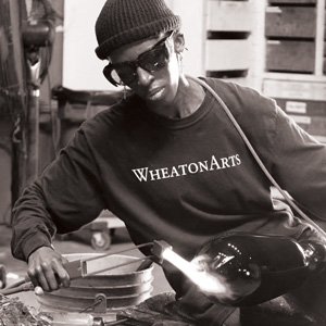 Grayscale image of Inga Lokwe working at a glassblowing bench and using a torch at the end of a long piece of hot glass that is at the end of a metal rod. There is a cord going across Inga's shoulder. Inga is wearing a beanie, dark glasses, and a dark long sleeve WheatonArts shirt.