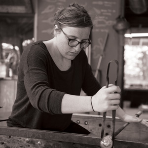 Grayscale image of Liesl Schubel sitting at a glassblowing bench. There is a metal rod across the sides of the bench and a small mass of hot glass at the end of it. Liesl is using a large pair of metal tweezers to shape the rod hot glass. Liesl has dark hair, glasses, and is looking towards the hot glass.